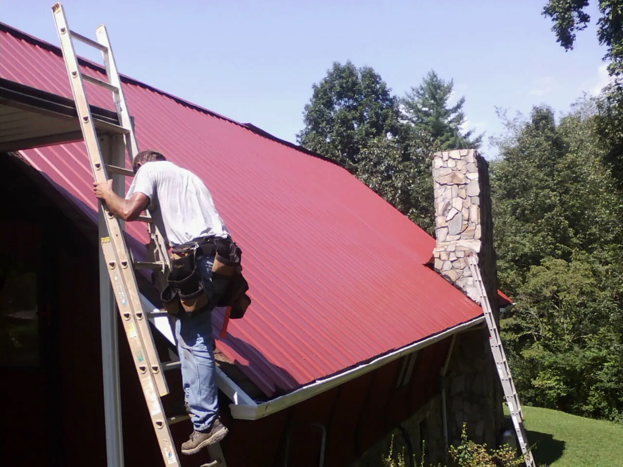 Roofer on a ladder beside a red metal roof and stone chimney — Soffit & Fascia Repair by Roofing Brothers in Moundsville